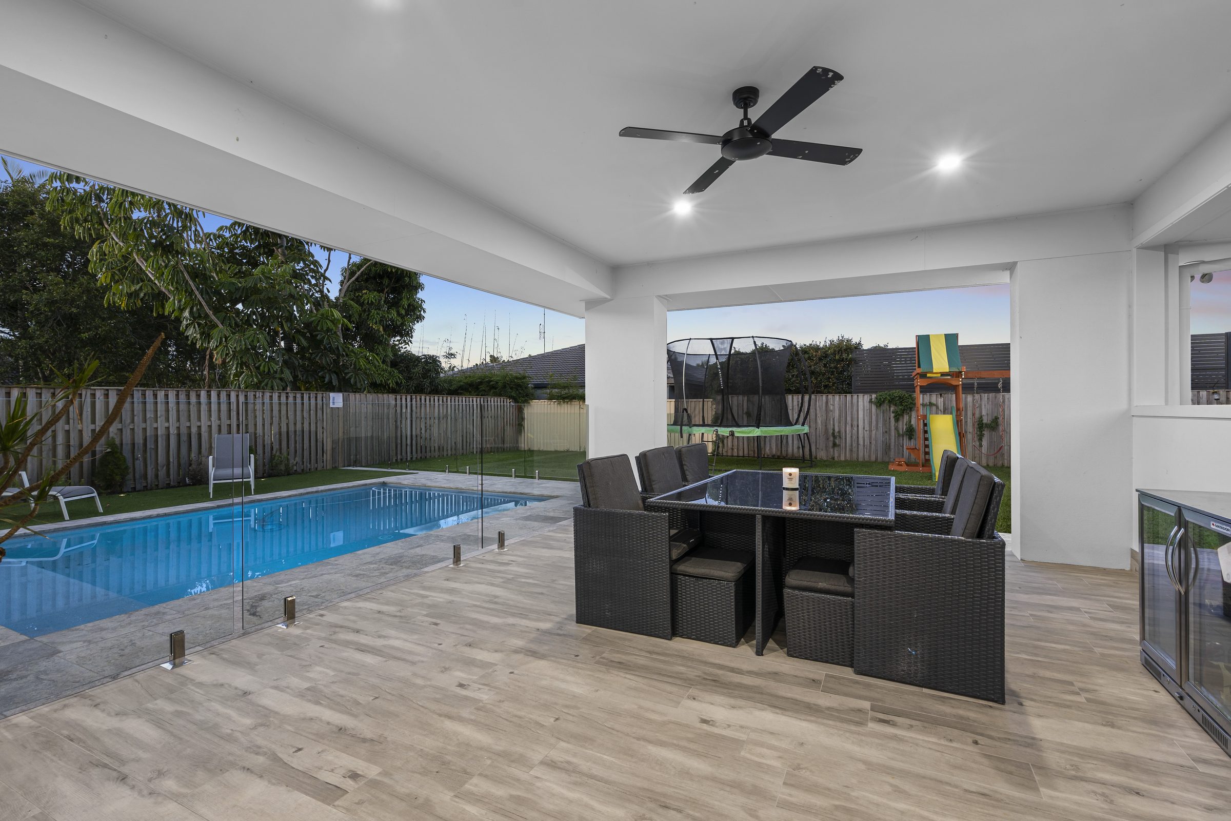Covered alfresco with an outdoor dining setting and ceiling fan, looking across a glass-fenced pool to the rear garden.