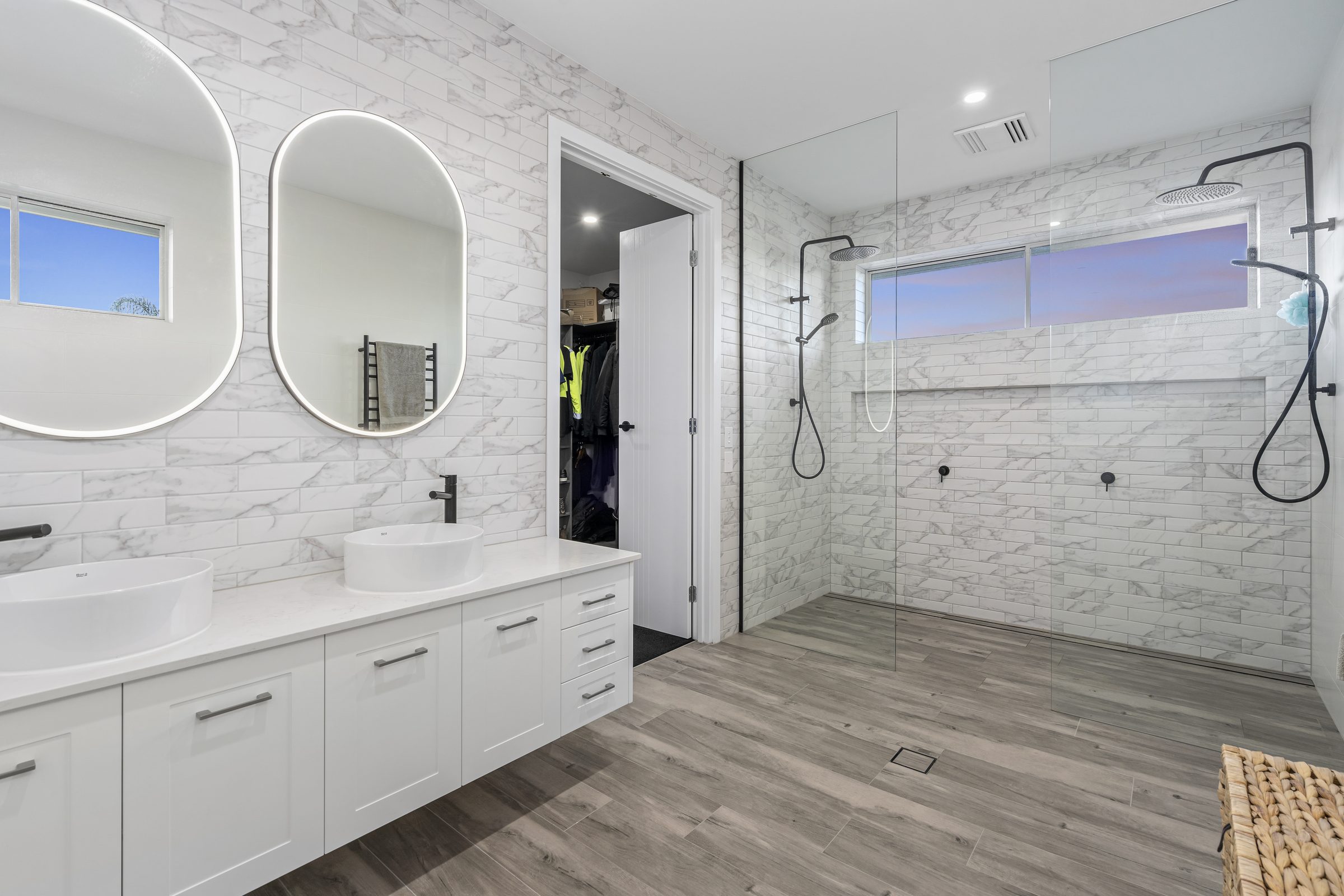 Main ensuite with a double vanity, two backlit oval mirrors, marble-look tiling and a walk-in shower alongside the robe.