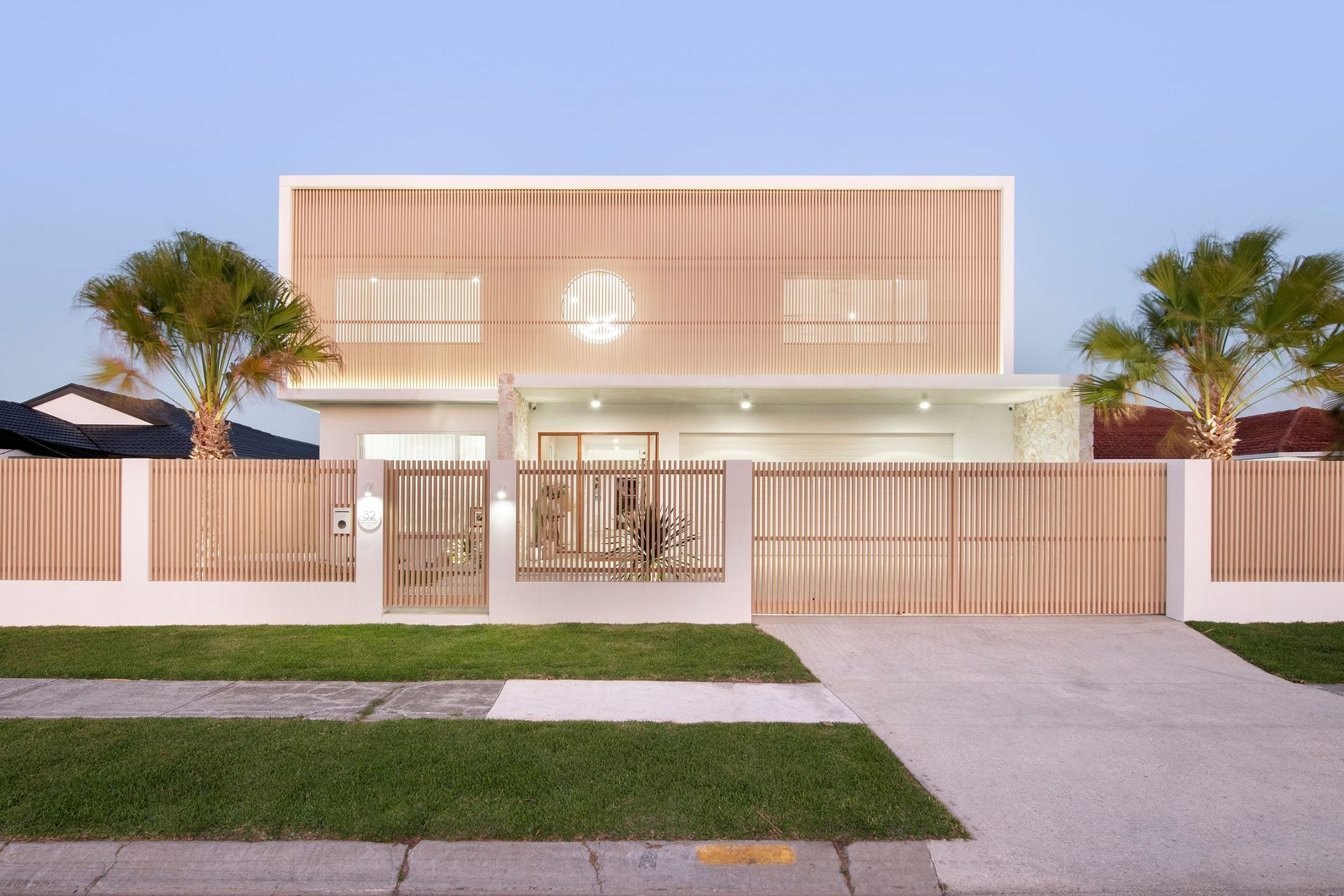 The street façade of the Village High Residence at dusk — a white rendered ground floor beneath a timber batten upper storey with a circular window at its centre, flanked by fan palms.