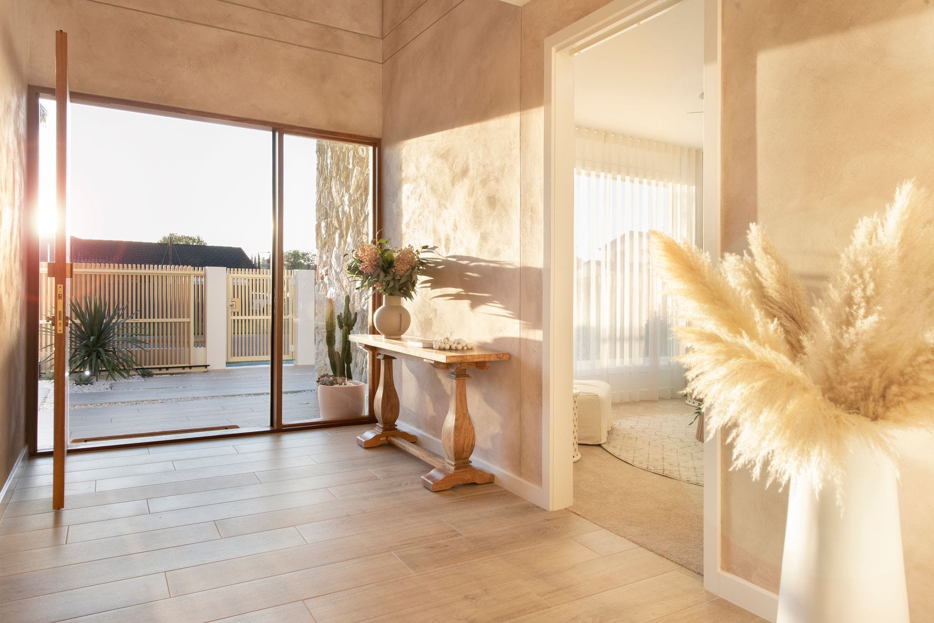 The entry hall in late afternoon light, with textured render, a stone feature wall, timber-look flooring and a console table beside full-height glazing onto the front courtyard.