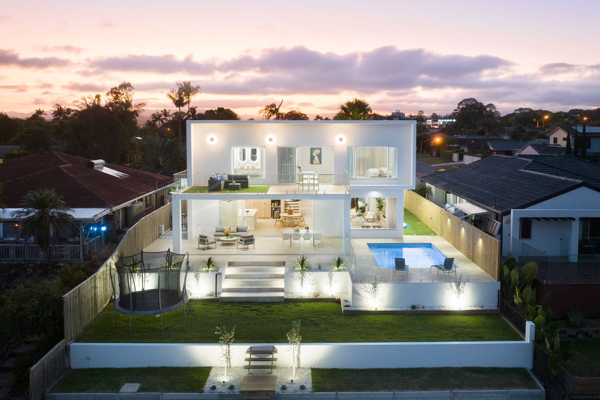 The rear elevation at dusk, lit from within, with the upper balcony set above a covered ground-floor terrace, the pool to one side and a stepped lawn below.